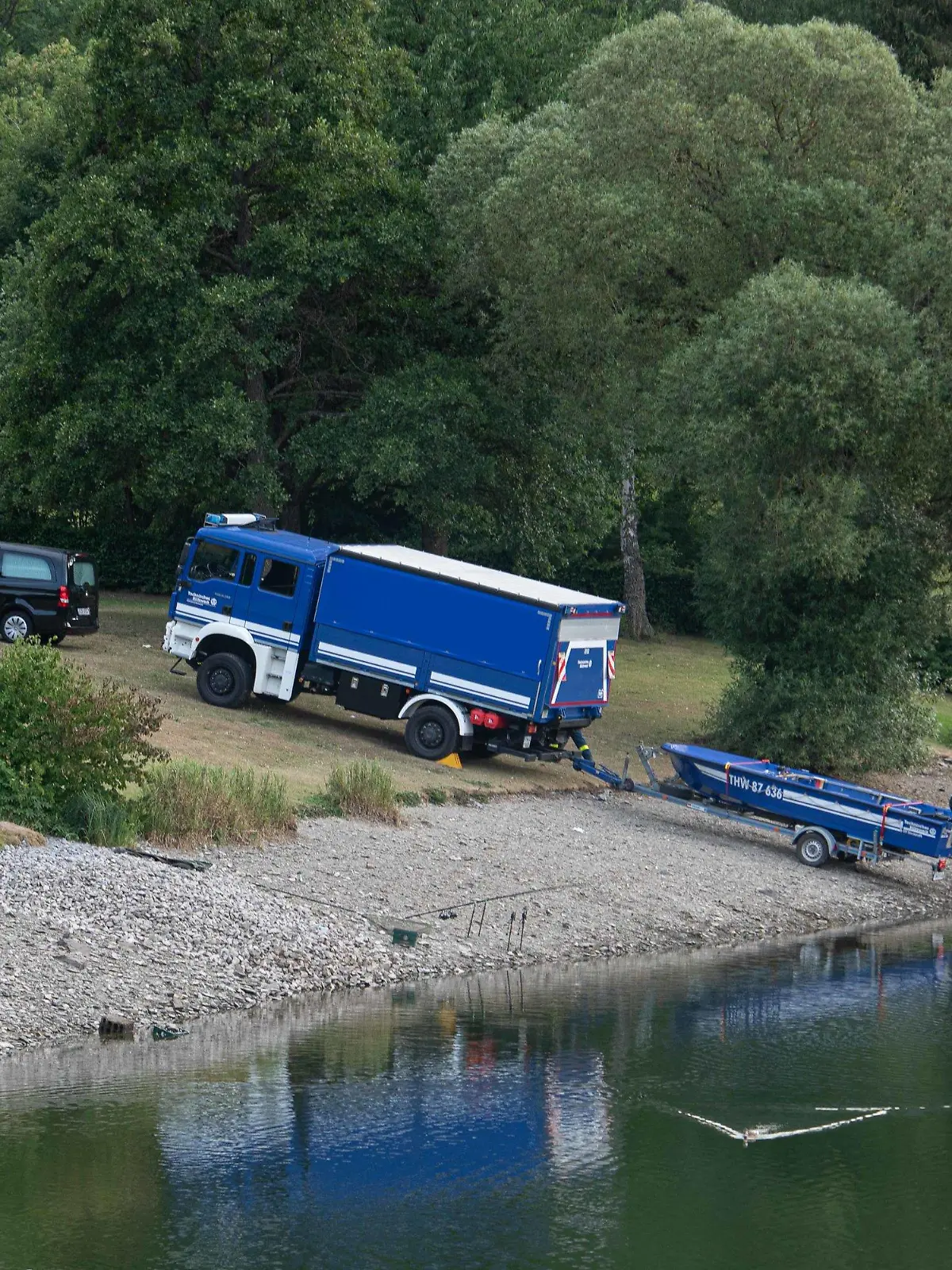 30.07.2022, Nordrhein-Westfalen, Simmerath: Ein Leichenwagen und ein Fahrzeug mit Boot des Technischen Hilfswerks (THW) stehen am Eiserbachsee. Ein 16 Jahre alter Junge ist in einem Badesee in Simmerath (Städteregion Aachen) untergegangen und gestorben. Der Jugendliche war am Samstagnachmittag in Begleitung eines Zehnjährigen in das Gewässer im Ortsteil Rurberg gesprungen, wie ein Polizeisprecher sagte. Beide waren offenbar Nichtschwimmer und drohten unterzugehen. Ersthelfern gelange es nach Angaben des Sprechers, das jüngere Kind zu retten. Der 16-Jährige wurde zwei Stunden später von Tauchern leblos aus dem Wasser geborgen. Foto: Vincent Kempf/dpa +++ dpa-Bildfunk +++