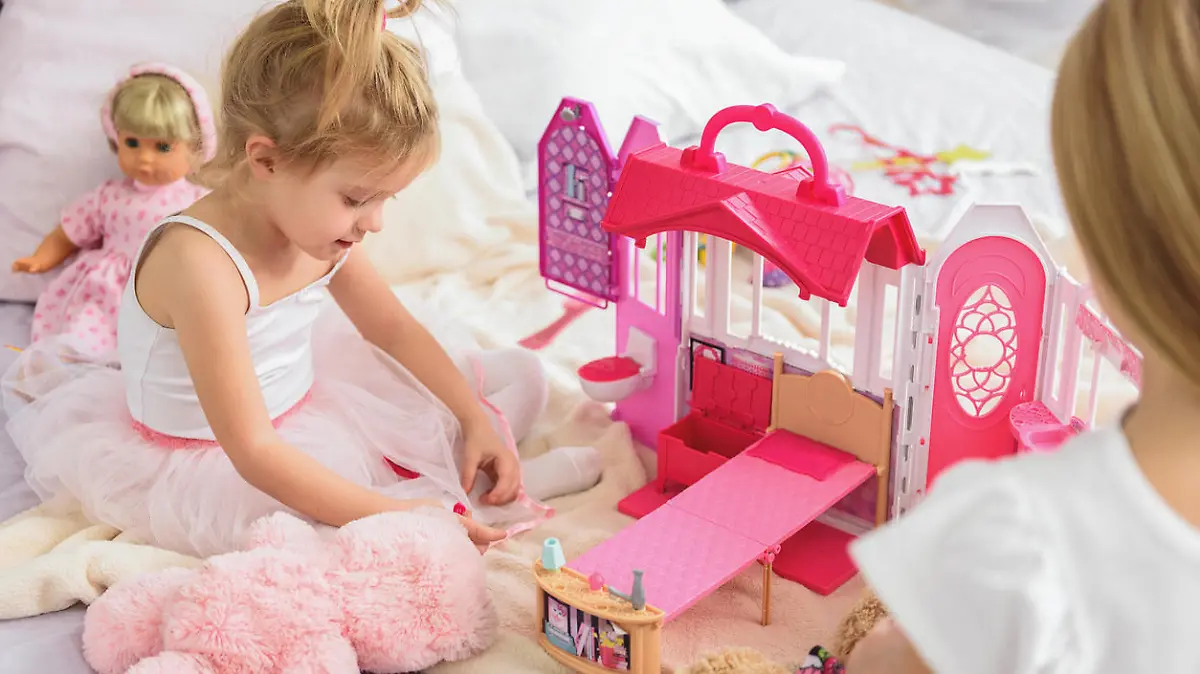 Pretty little child is enjoying game in doll house with her elder sister. She is sitting on bed and smiling