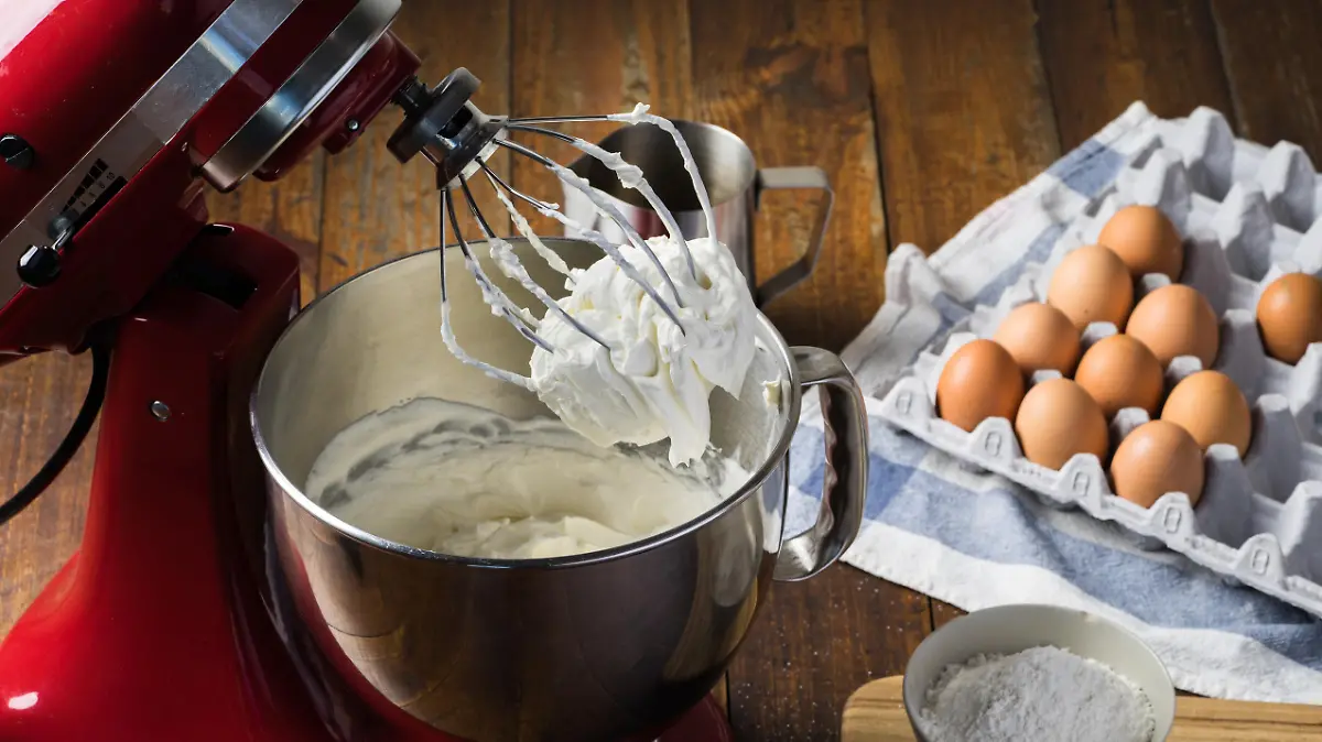 Standing mixer with cream on whisk on wooden table with ingredients.