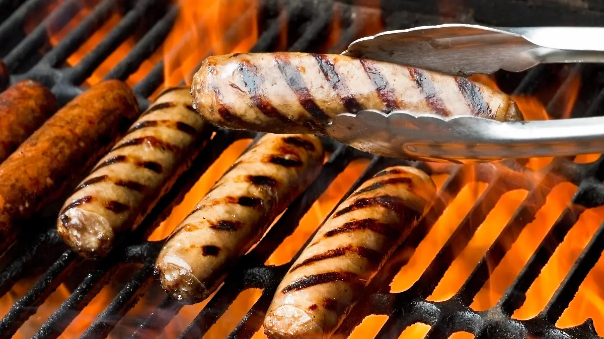 Bratwurst, hot dogs or polish sausage on a flaming charcoal grill.  One brat is being presented in a pair of tongs in front of the others.  All of them have beautiful appetizing grill marks as the flames kiss the meat sealing in the flavors.