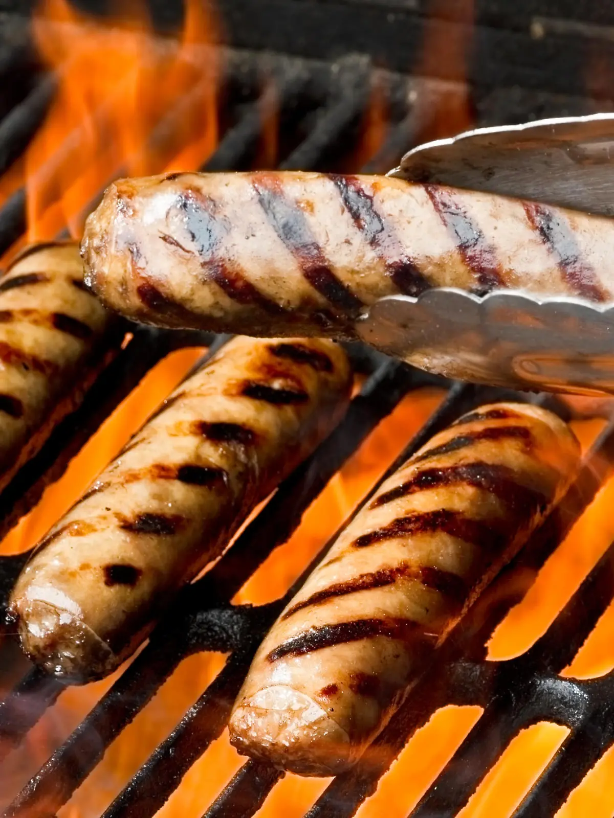 Bratwurst, hot dogs or polish sausage on a flaming charcoal grill.  One brat is being presented in a pair of tongs in front of the others.  All of them have beautiful appetizing grill marks as the flames kiss the meat sealing in the flavors.