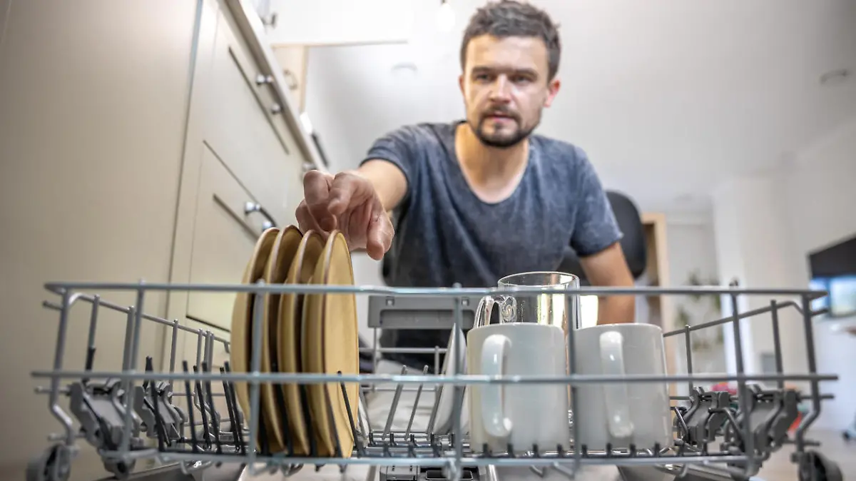 A man in front of an open dishwasher takes out or puts down dishes.