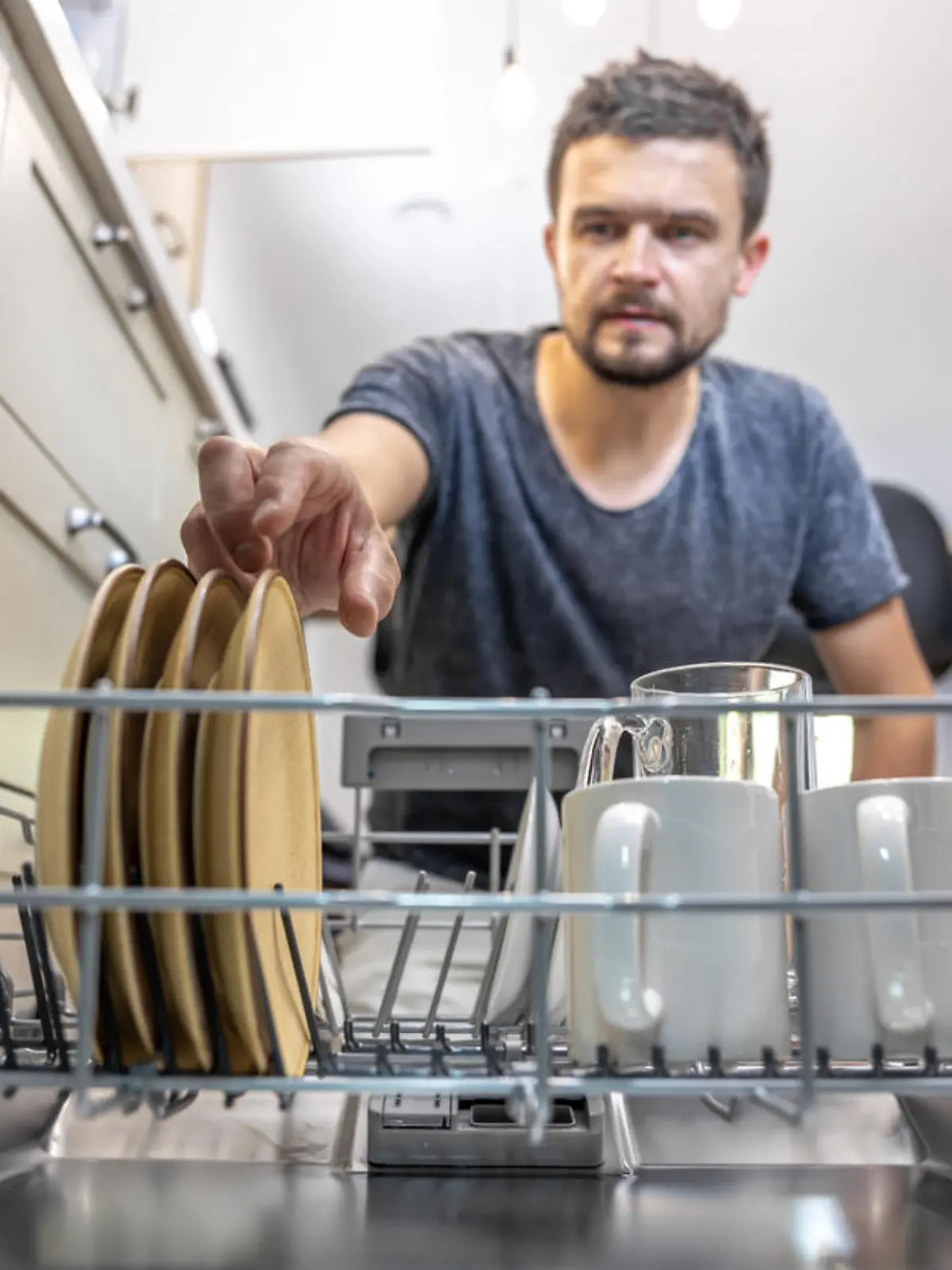 A man in front of an open dishwasher takes out or puts down dishes.