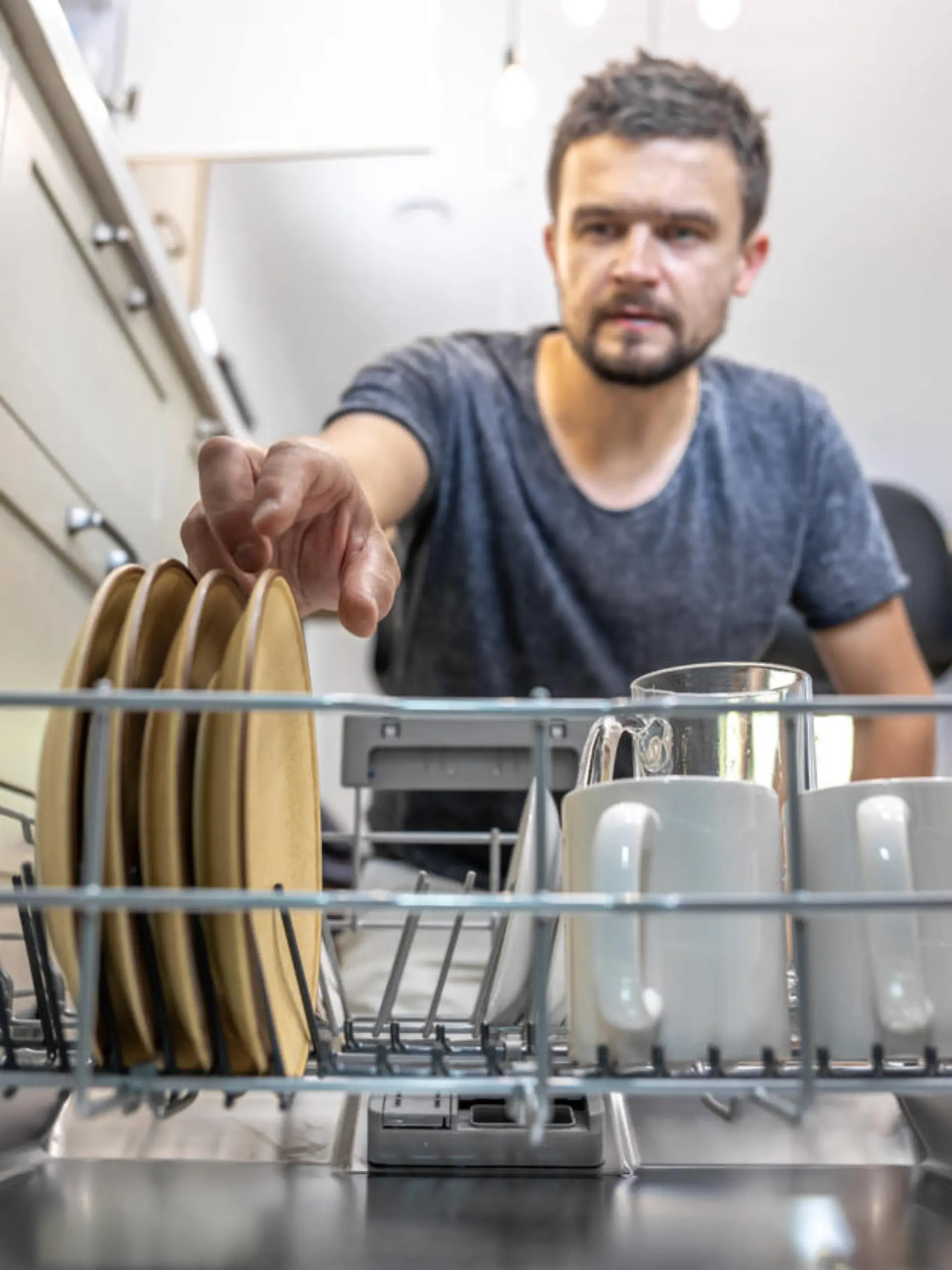 A man in front of an open dishwasher takes out or puts down dishes.