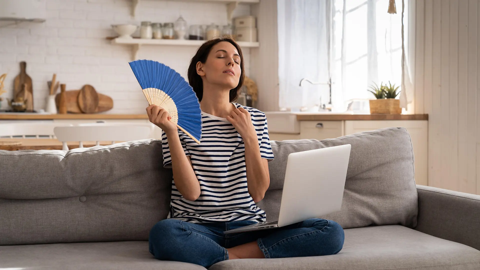 Tired millennial woman suffers from stuffiness and an inoperative air conditioner, waving blue fan sitting on couch at home working on laptop computer. Overheating high temperature, hot summer weather