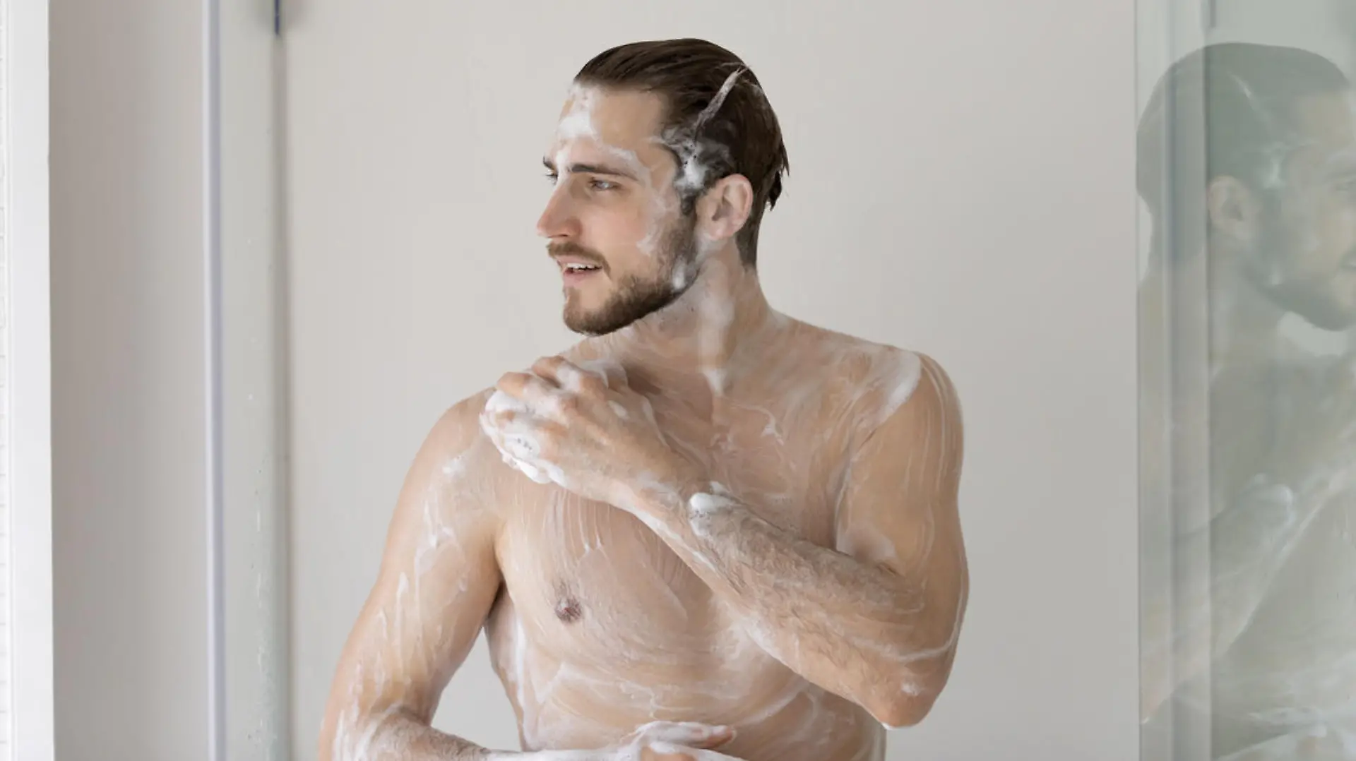 Smiling young man standing in bathroom, taking shower, washing body with natural moisturizing gel and hair with shampoo, enjoying morning routine, personal hygiene and skincare concept