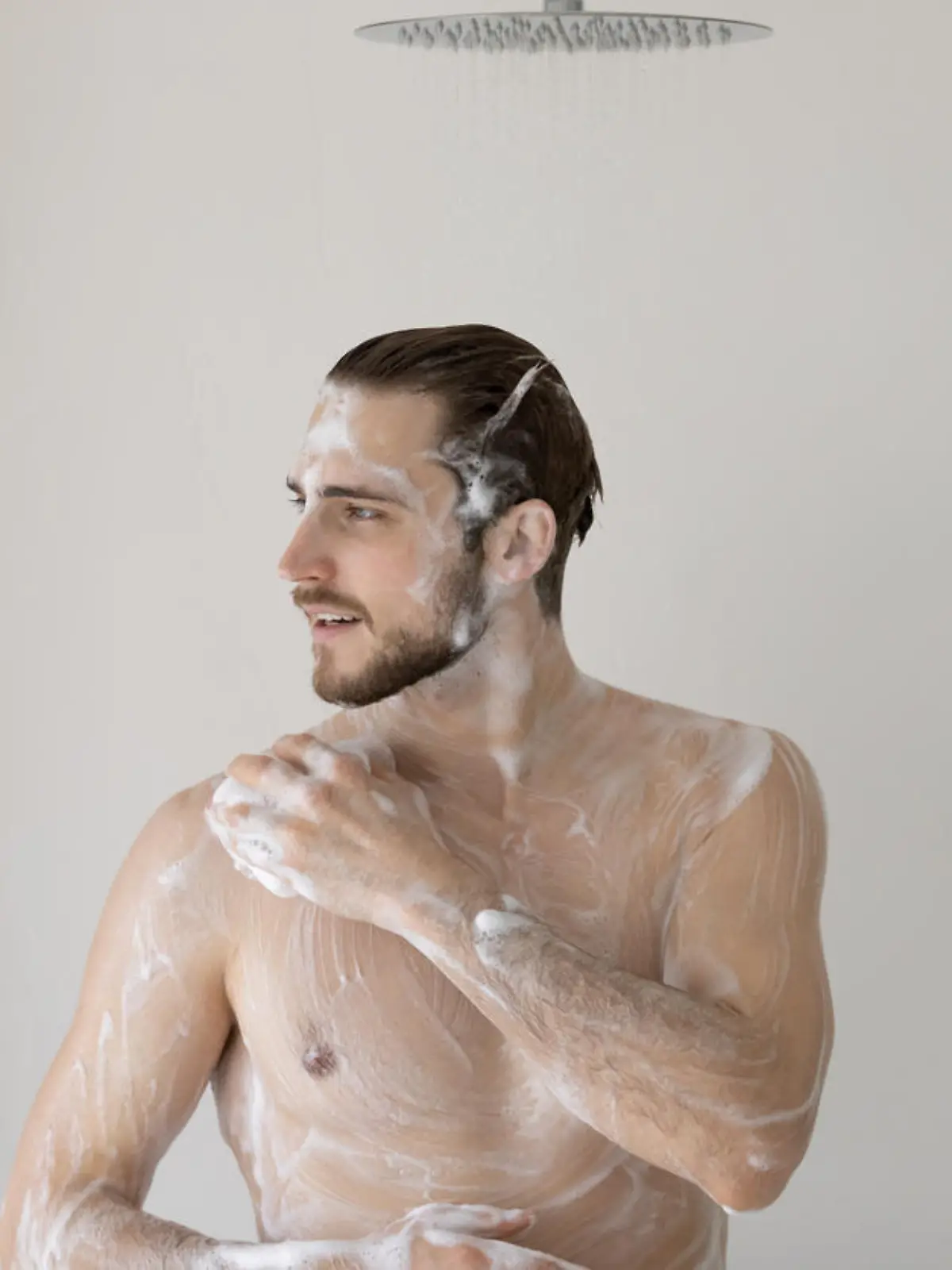 Smiling young man standing in bathroom, taking shower, washing body with natural moisturizing gel and hair with shampoo, enjoying morning routine, personal hygiene and skincare concept