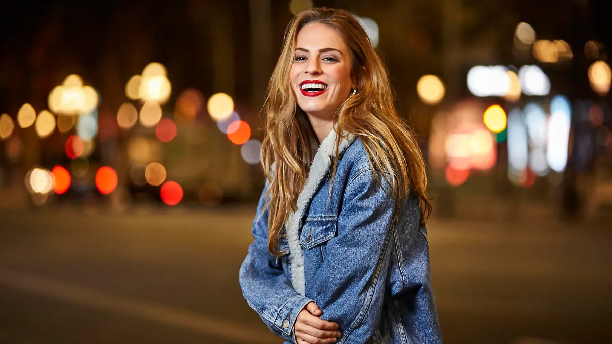 Portrait of smiling young woman standing on street. Cheerful female with brown hair wearing denim jacket. She is in city at night.