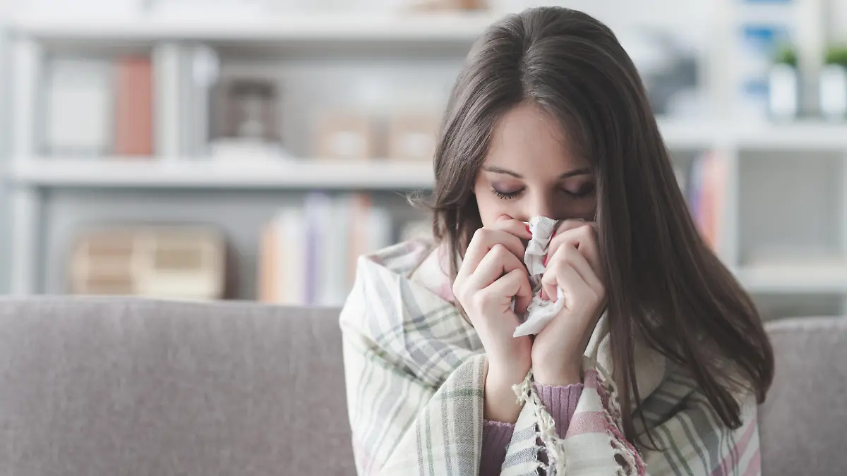 Sick young woman at home on the sofa with a cold, she is covering with a blanket and blowing her nose