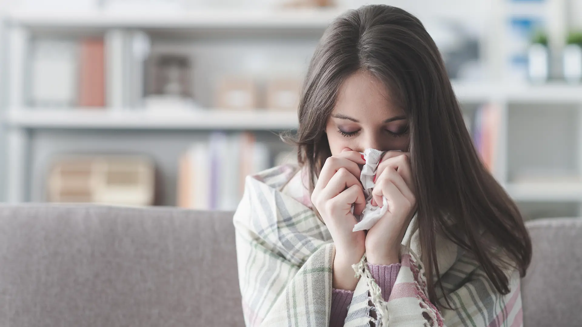 Sick young woman at home on the sofa with a cold, she is covering with a blanket and blowing her nose