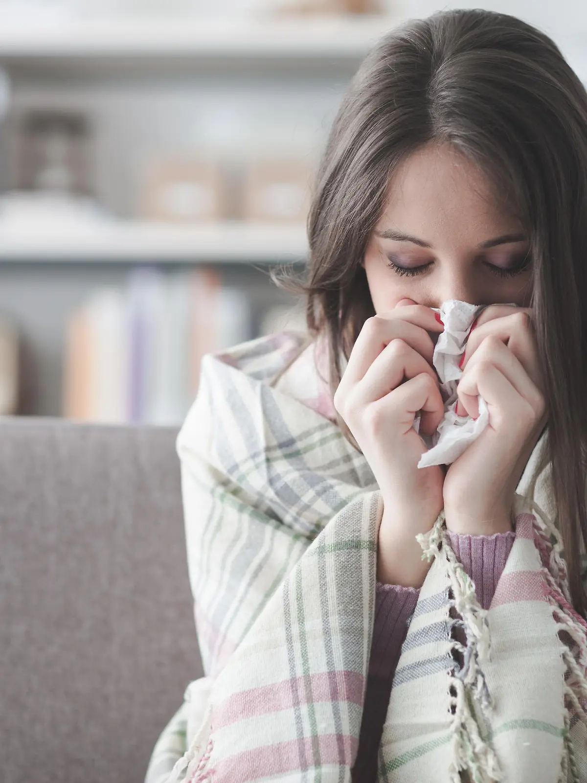 Sick young woman at home on the sofa with a cold, she is covering with a blanket and blowing her nose