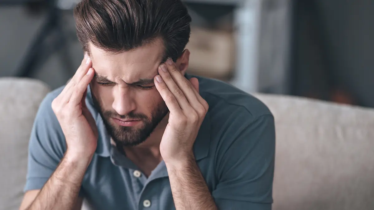 Frustrated handsome young man touching his head and keeping eyes closed while sitting on the couch at home
