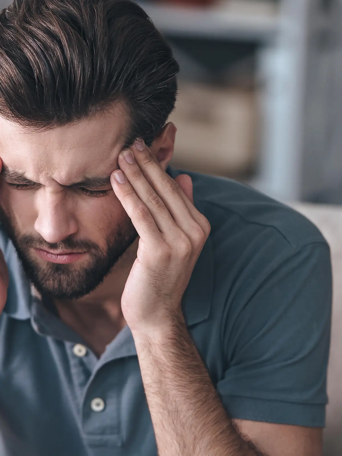 Frustrated handsome young man touching his head and keeping eyes closed while sitting on the couch at home