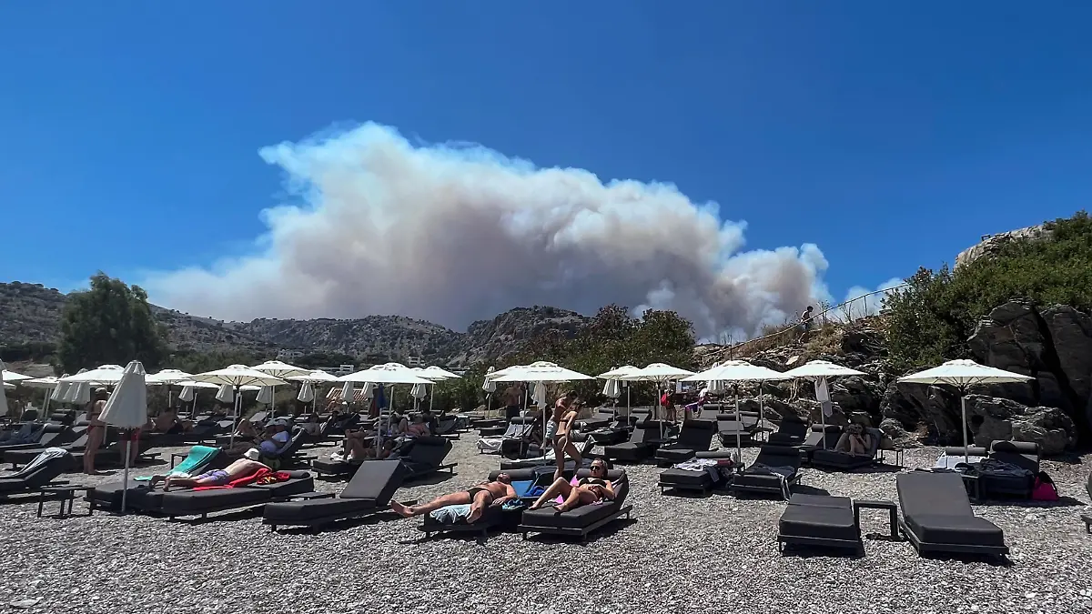 22.07.2023, Griechenland, Lindos: Am Viglika Beach auf der griechischen Insel Lindos aus, wo sich Menschen auf Sonnenliegen am Strand aufhalten, kann man aufsteigenden Rauch von Waldbränden auf der Ferieninsel Rhodos sehen. Waldbrände werden aufgrund starker Winde wieder stärker. Es wird erwartet, dass sich die Hitze in Griechenland im Laufe des Wochenendes verstärkt und sich 44 Grad Celsius nähert. Das ganze Land soll bis Ende Juli eine weitere Hitzewelle erleben. Foto: Mark Cosgrove/News Images via ZUMA Press Wire/dpa +++ dpa-Bildfunk +++