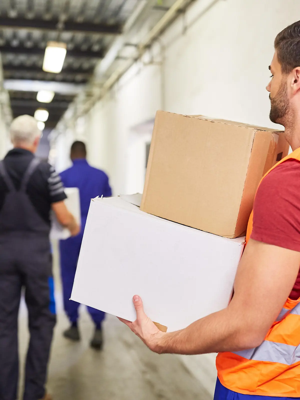 Arbeiter mit Warnweste trägt Kartons durch Gang zum Lager einer Fabrik || Modellfreigabe vorhanden