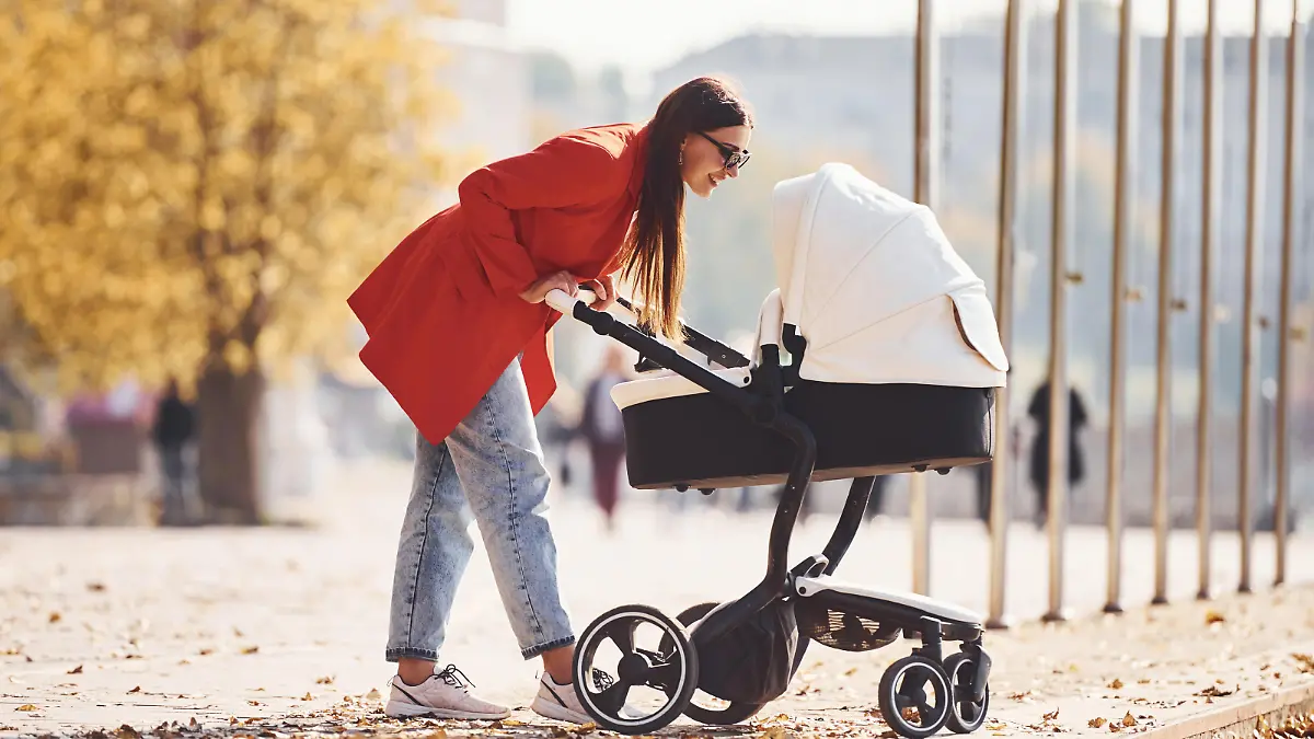 Mother in red coat have a walk with her kid in the pram in the park at autumn time.