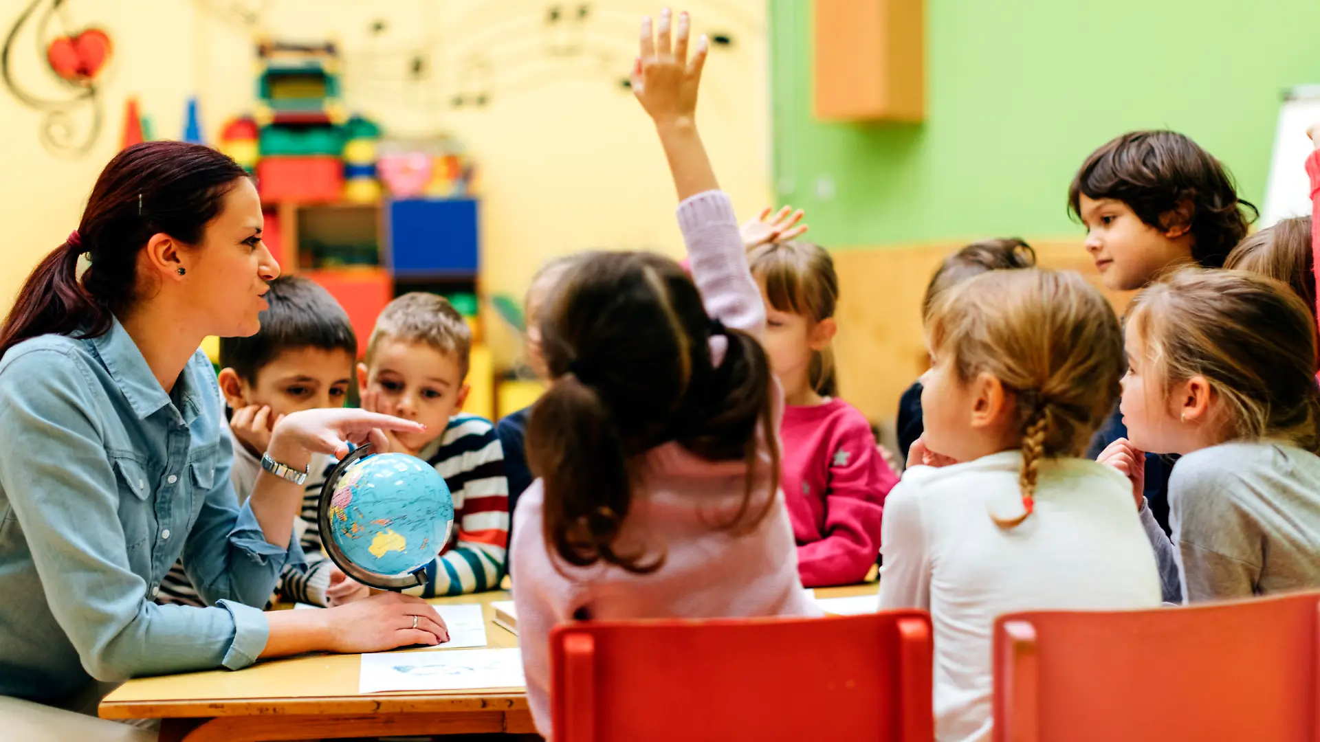 Preschool teacher teaching her children about geography. Using globe and asking the questions. Children answering the questions. They are sitting by the table. Models in this shot are part of real kindergarten group and their teacher.