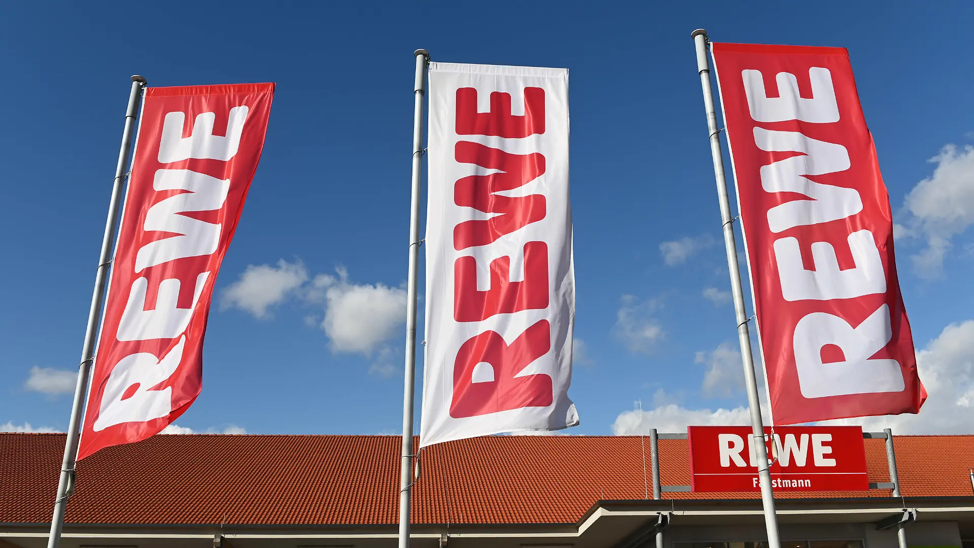 ARCHIV - 25.02.2020, Hessen, Volkmarsen: Fahnen mit dem Logo des Lebensmitteleinzelhändlers Rewe wehen vor einer Filiale. Die Supermarktkette Rewe verzichtet künftig auf ihre Prospekte. (zu dpa «Rewe stellt Werbeprospekte ein») Foto: Uwe Zucchi/dpa +++ dpa-Bildfunk +++