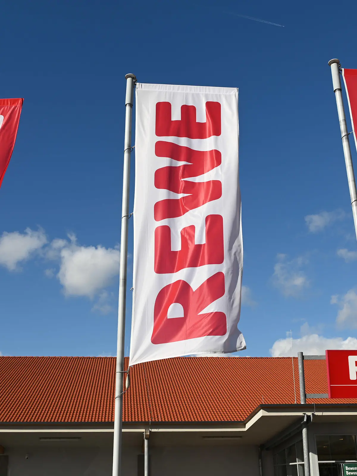 ARCHIV - 25.02.2020, Hessen, Volkmarsen: Fahnen mit dem Logo des Lebensmitteleinzelhändlers Rewe wehen vor einer Filiale. Die Supermarktkette Rewe verzichtet künftig auf ihre Prospekte. (zu dpa «Rewe stellt Werbeprospekte ein») Foto: Uwe Zucchi/dpa +++ dpa-Bildfunk +++