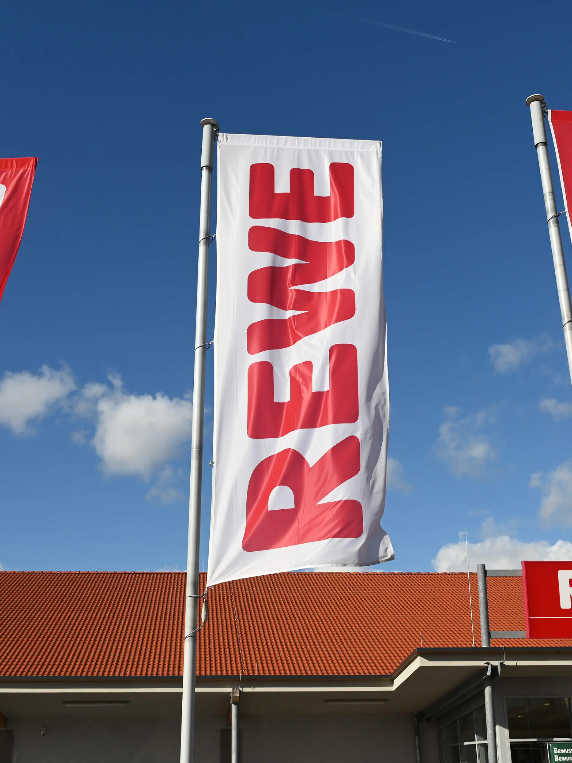 ARCHIV - 25.02.2020, Hessen, Volkmarsen: Fahnen mit dem Logo des Lebensmitteleinzelhändlers Rewe wehen vor einer Filiale. Die Supermarktkette Rewe verzichtet künftig auf ihre Prospekte. (zu dpa «Rewe stellt Werbeprospekte ein») Foto: Uwe Zucchi/dpa +++ dpa-Bildfunk +++