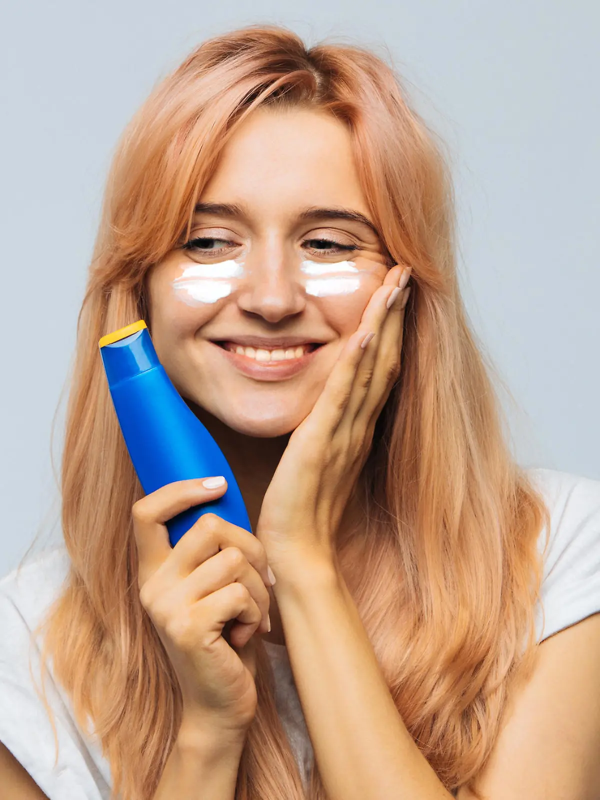 Portrait of young joyful cute woman smiling and applying suntan cream (sunscreen lotion) on face. Pretty female smeared face, cheeks with sun protection cream. Sunburn, vacation, sun concept