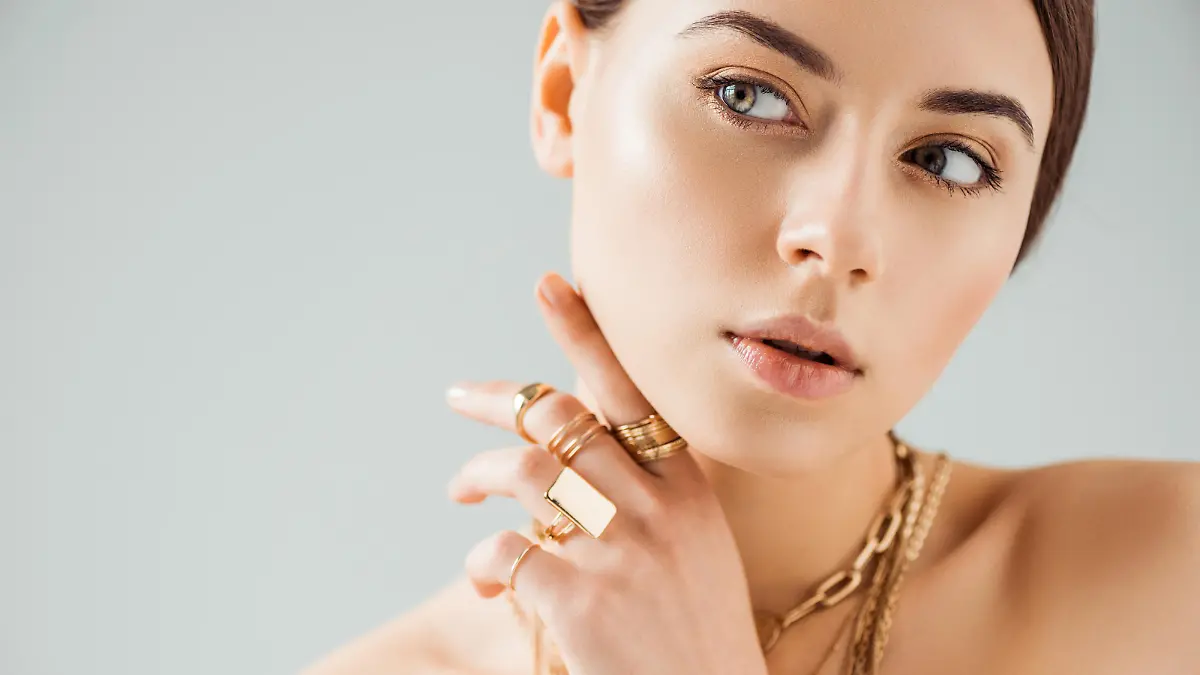 young nude woman in golden rings and necklaces looking away isolated on grey