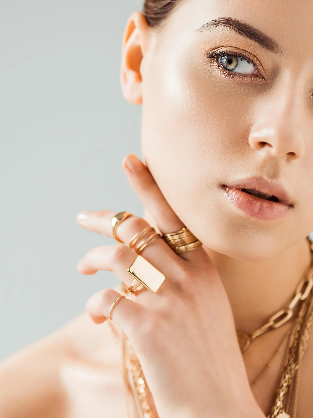 young nude woman in golden rings and necklaces looking away isolated on grey