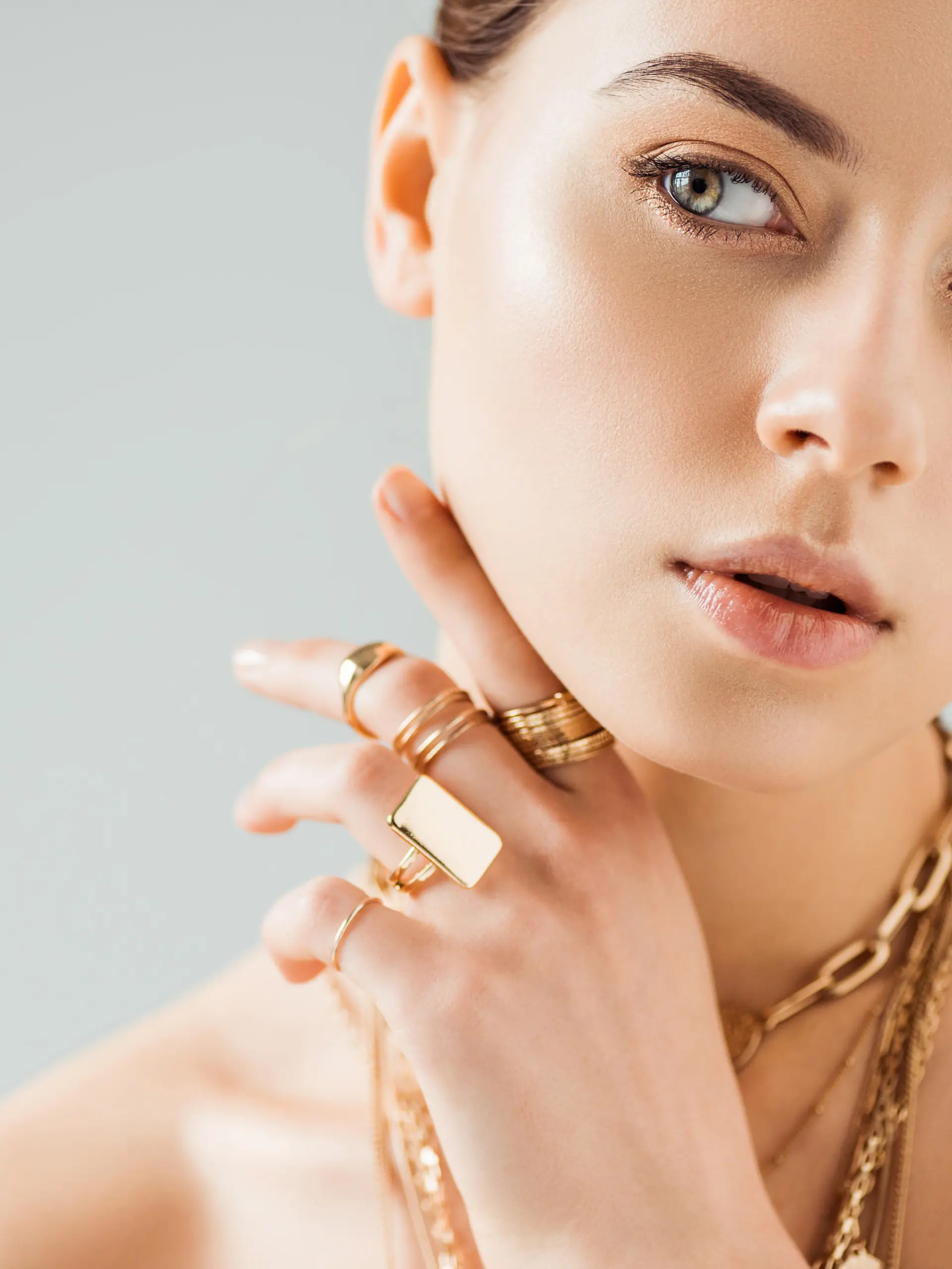 young nude woman in golden rings and necklaces looking away isolated on grey