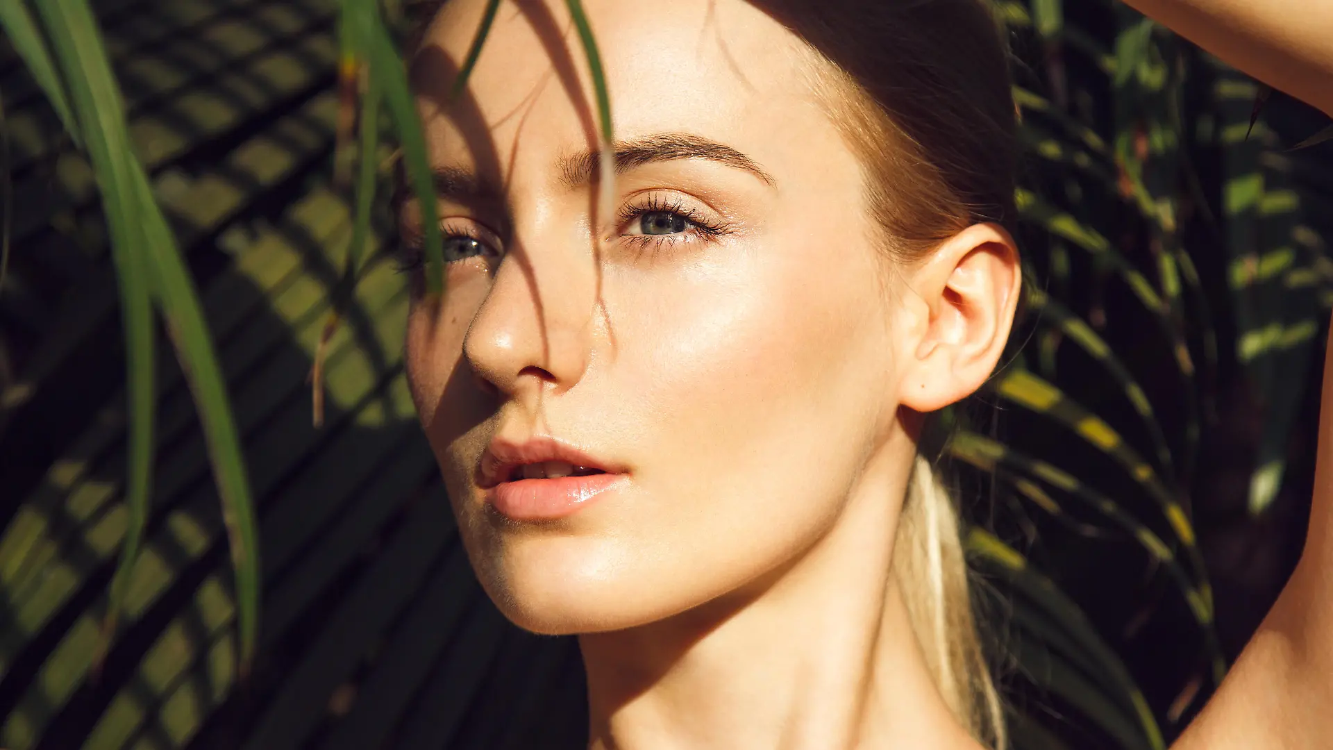 Close-up portrait of beautiful blonde young woman with perfect skin, beautiful green eyes and long neck hiding under palm leaf