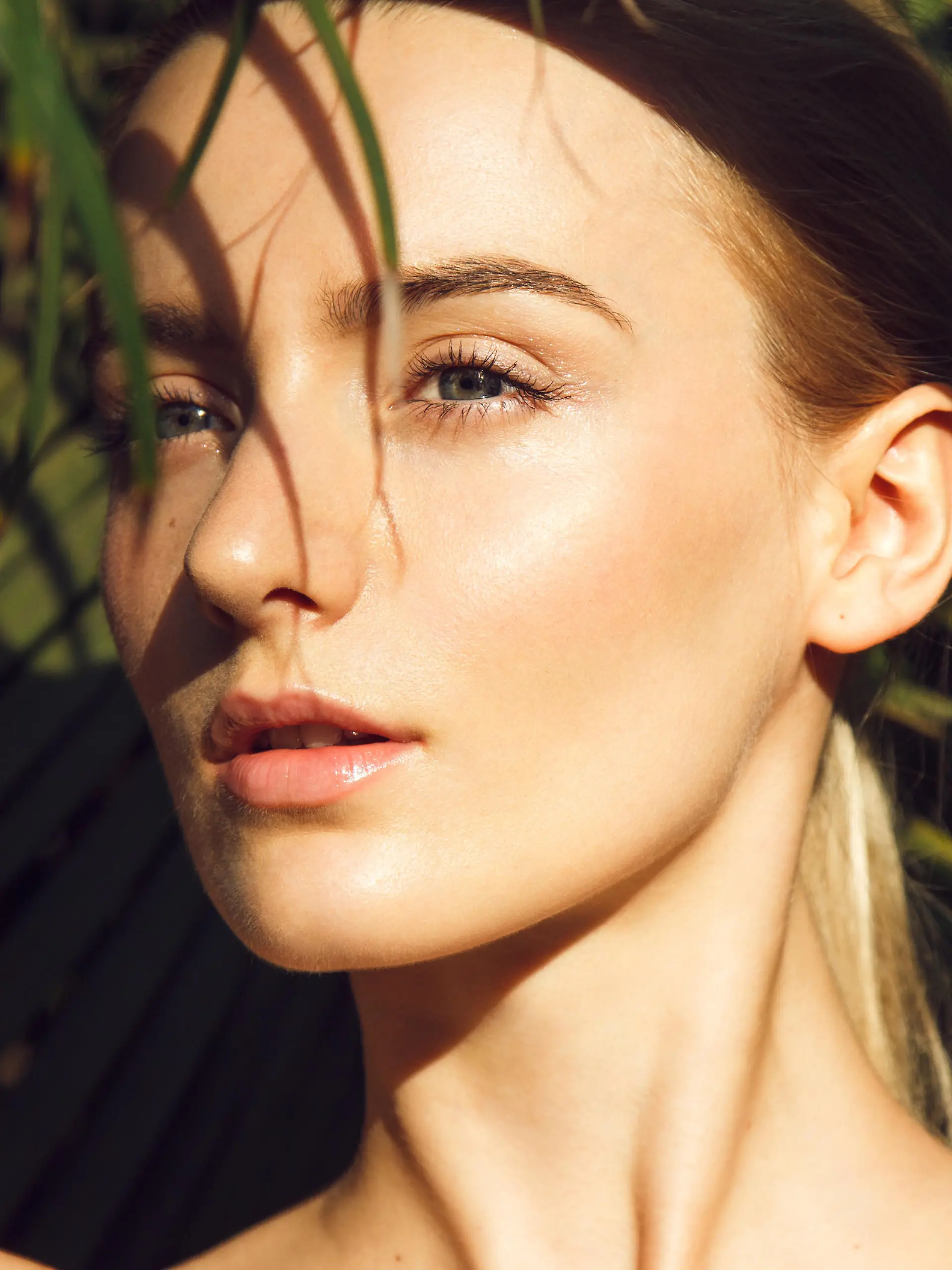 Close-up portrait of beautiful blonde young woman with perfect skin, beautiful green eyes and long neck hiding under palm leaf
