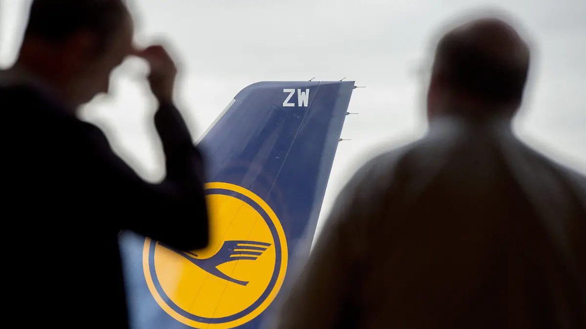 ARCHIV - 07.11.2015, Hessen, Frankfurt/Main: Zwei Männer blicken auf dem Flughafen durch ein Fenster auf eine abgestellte Lufthansa-Maschine. Lufthansa und die Gewerkschaften ringen weiter um ein Sparpaket für die angeschlagene Fluggesellschaft. Tausende Stellen stehen auf der Kippe. (Zu dpa «Tausende Stellen bei Lufthansa auf der Kippe - Ringen um Sparpaket») Foto: Boris Roessler/dpa +++ dpa-Bildfunk +++