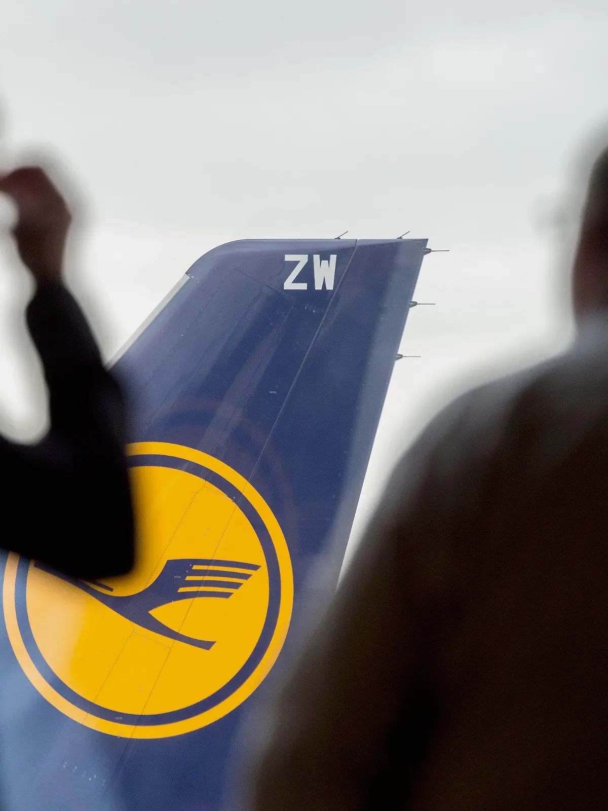 ARCHIV - 07.11.2015, Hessen, Frankfurt/Main: Zwei Männer blicken auf dem Flughafen durch ein Fenster auf eine abgestellte Lufthansa-Maschine. Lufthansa und die Gewerkschaften ringen weiter um ein Sparpaket für die angeschlagene Fluggesellschaft. Tausende Stellen stehen auf der Kippe. (Zu dpa «Tausende Stellen bei Lufthansa auf der Kippe - Ringen um Sparpaket») Foto: Boris Roessler/dpa +++ dpa-Bildfunk +++