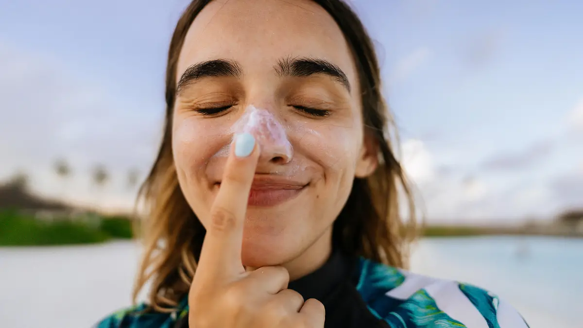 Photo of a young woman applying sunscreen while being outdoors, at the beach; making silly faces while reapplying SPF and protecting her skin from the sun.