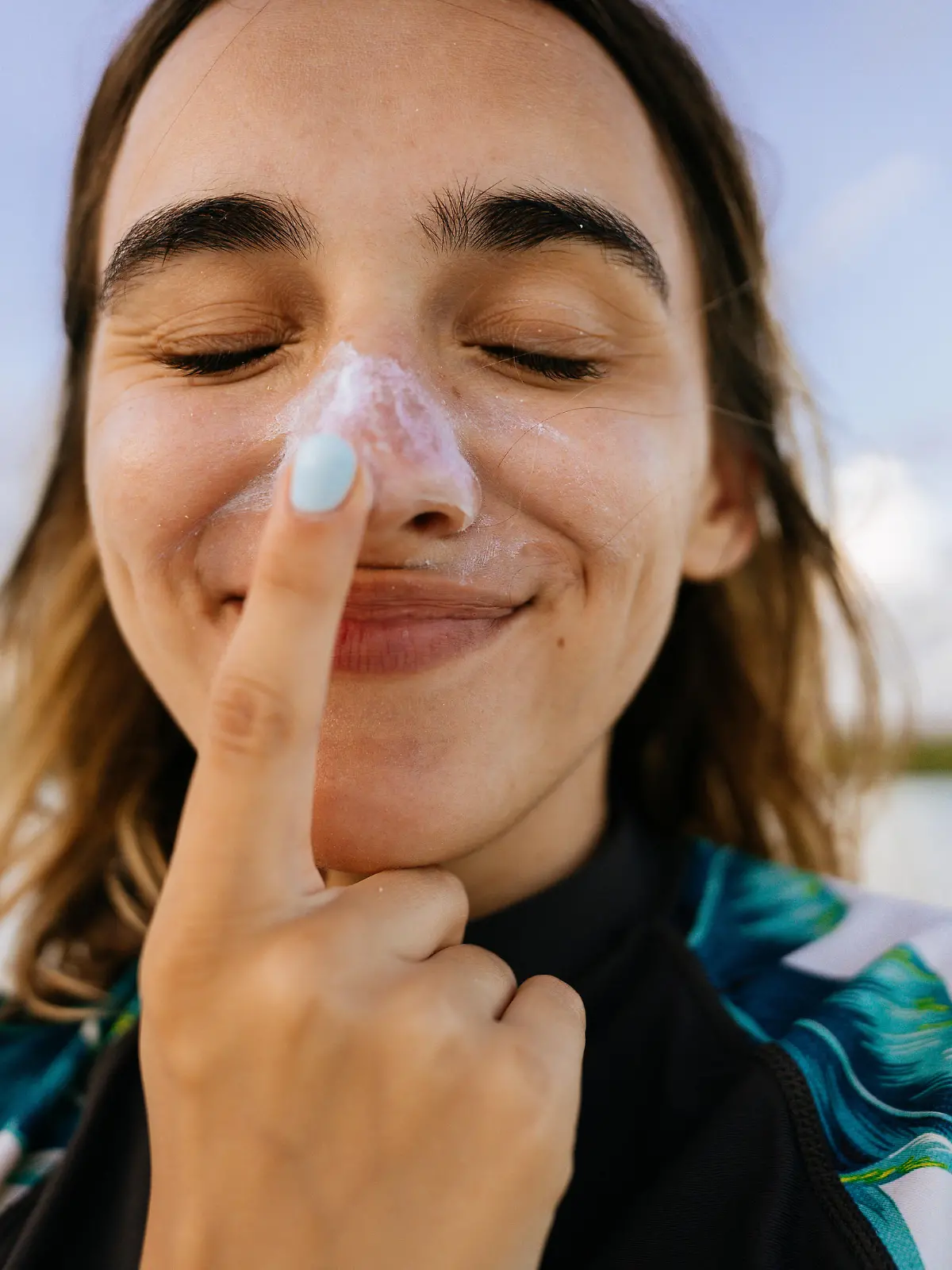 Photo of a young woman applying sunscreen while being outdoors, at the beach; making silly faces while reapplying SPF and protecting her skin from the sun.