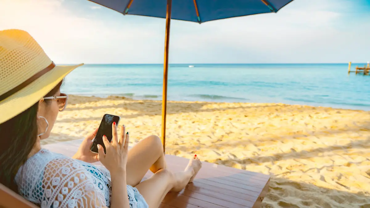 Asian woman with hat sit on sunbed under beach umbrella at sand beach and using smartphone on summer vacation. Girl in casual style relax and enjoy holiday at tropical paradise beach. Summer vibes.