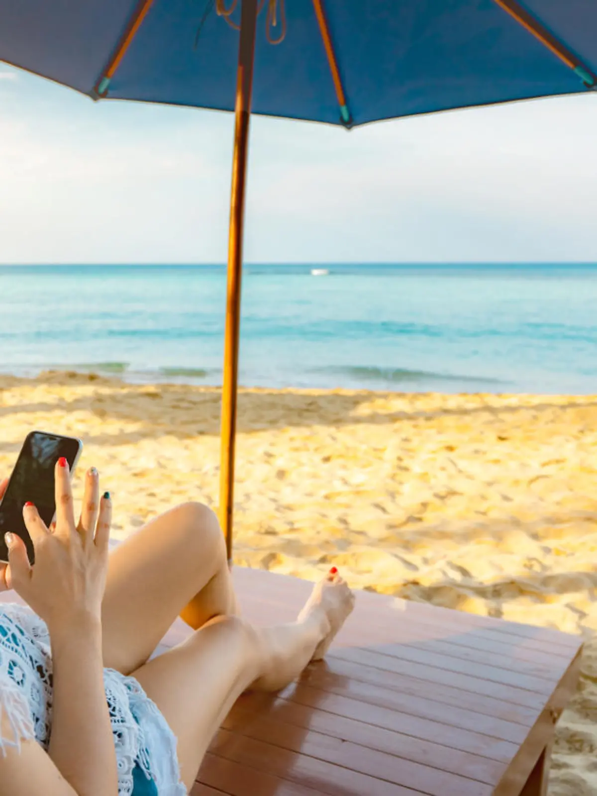 Asian woman with hat sit on sunbed under beach umbrella at sand beach and using smartphone on summer vacation. Girl in casual style relax and enjoy holiday at tropical paradise beach. Summer vibes.