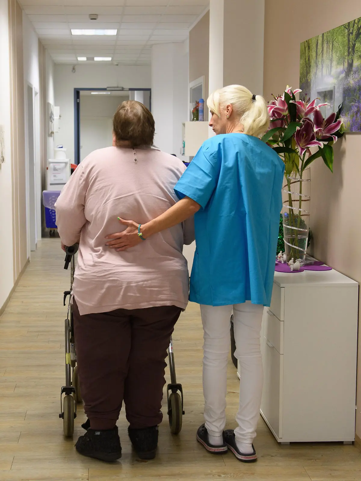 Pflegerin und Patientin mit Rollator bei einem Spaziergang über den Flur der Pflegeeinrichtung.