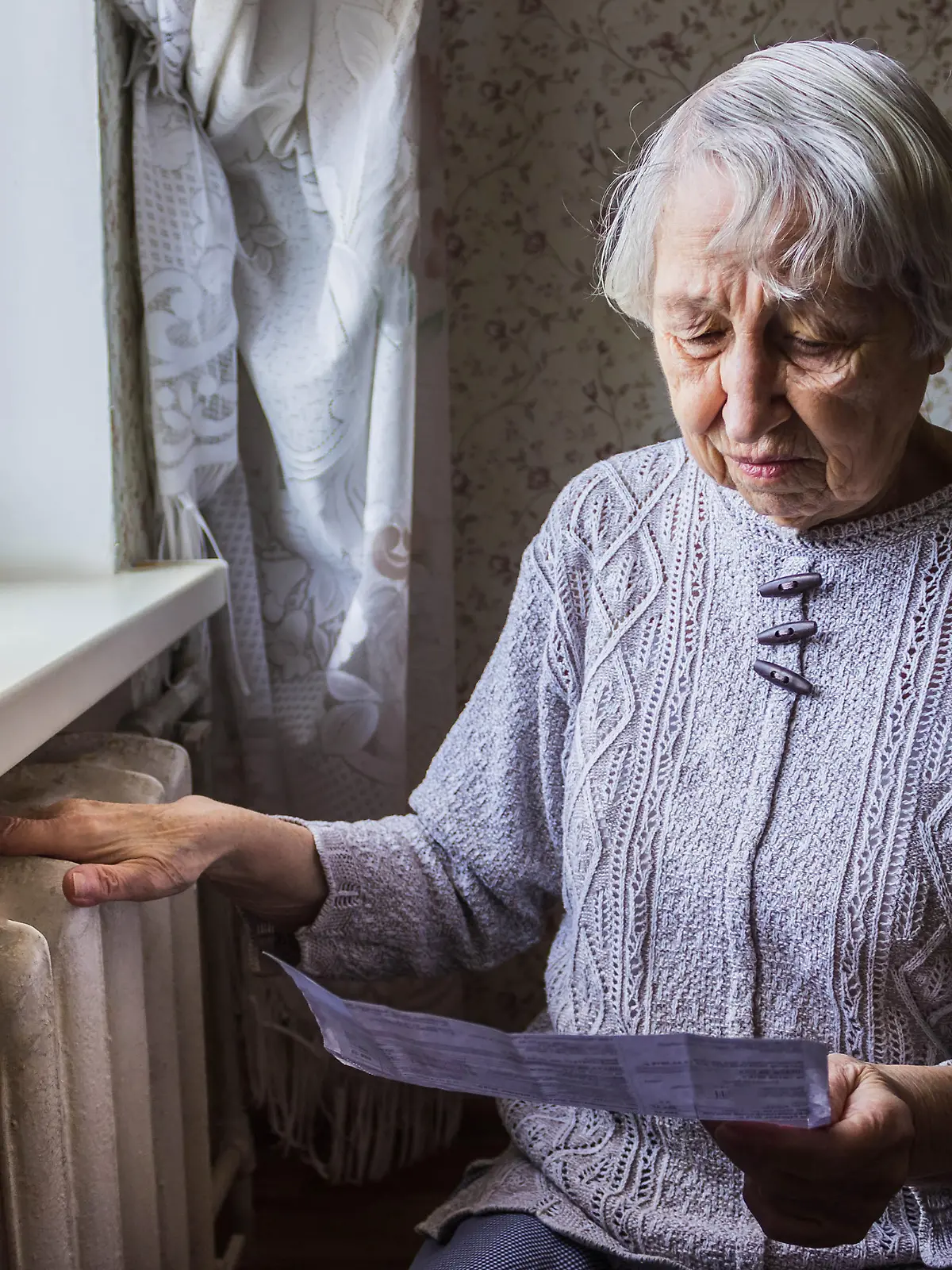 The senior woman holding gas bill in front of heating radiator. Payment for heating in winter.