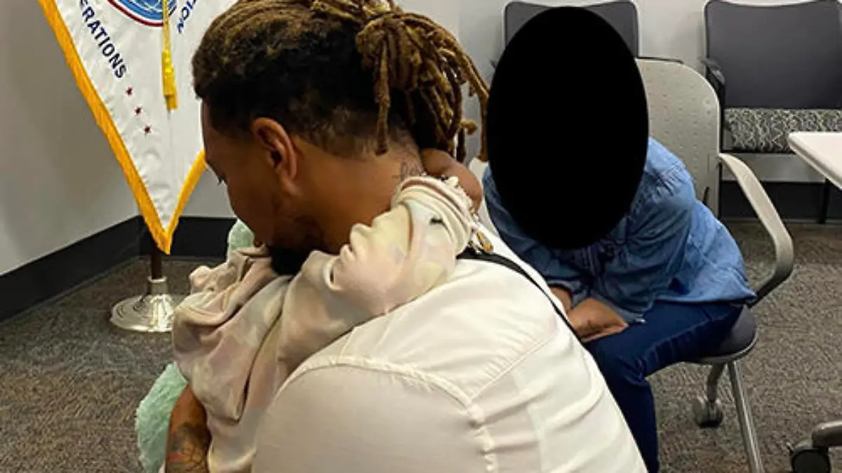 Image of Majesty and her father hugging, you mostly see him- a black man with brown and blonde dreads tied back hugs his daughter so you can just see her arms around his neck and they sit in a Federal office with a field opperations flag of white and blue in the background.