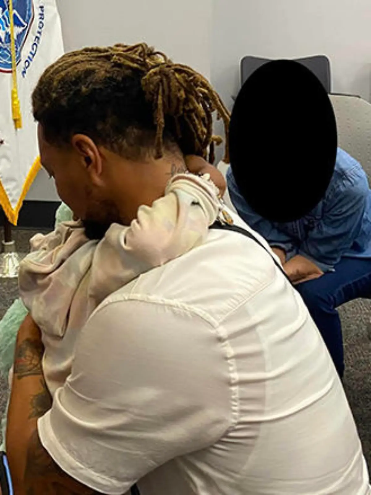 Image of Majesty and her father hugging, you mostly see him- a black man with brown and blonde dreads tied back hugs his daughter so you can just see her arms around his neck and they sit in a Federal office with a field opperations flag of white and blue in the background.