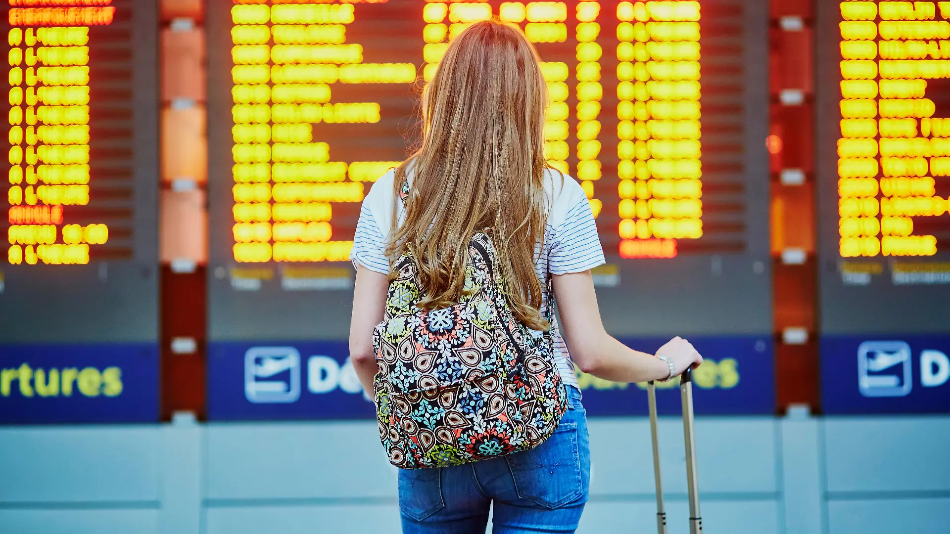 Beautiful young tourist girl with backpack and carry on luggage in international airport, near flight information board