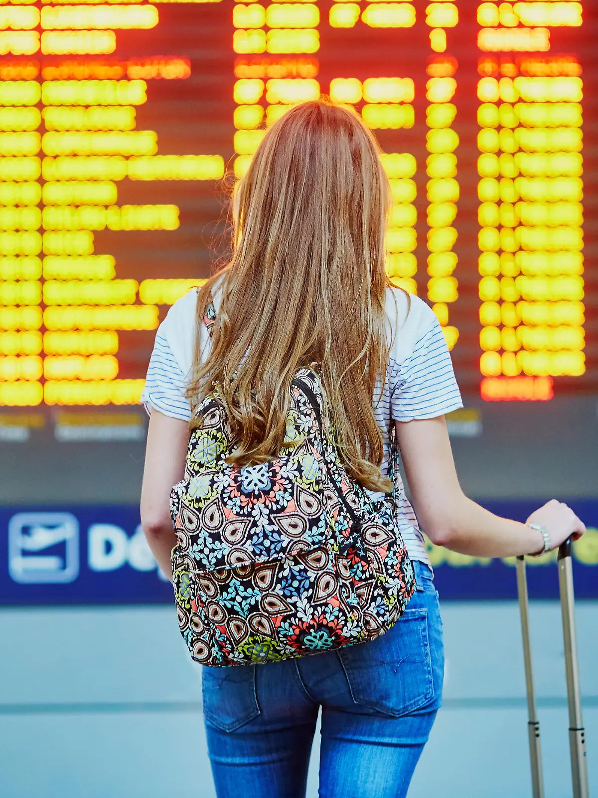 Beautiful young tourist girl with backpack and carry on luggage in international airport, near flight information board
