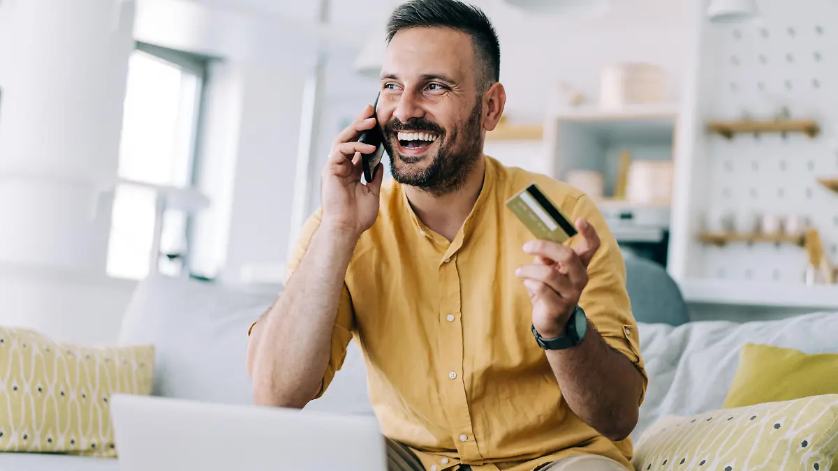 Shot of handsome young man shopping online using his laptop and mobile phone while sitting on the sofa at home. Smiling young businessman holding a credit card and using a laptop and smartphone at home.