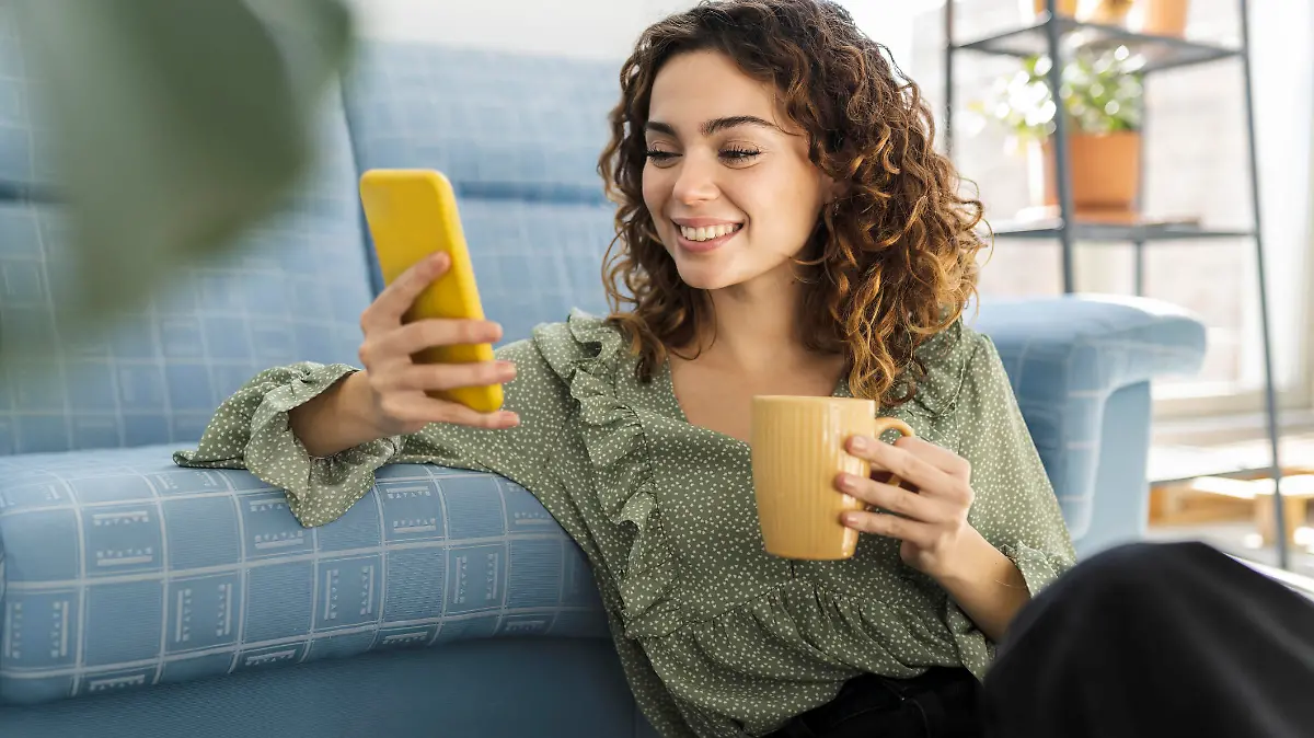 curly haired woman sitting on sofa with a cup of hot drink and mobile phone
