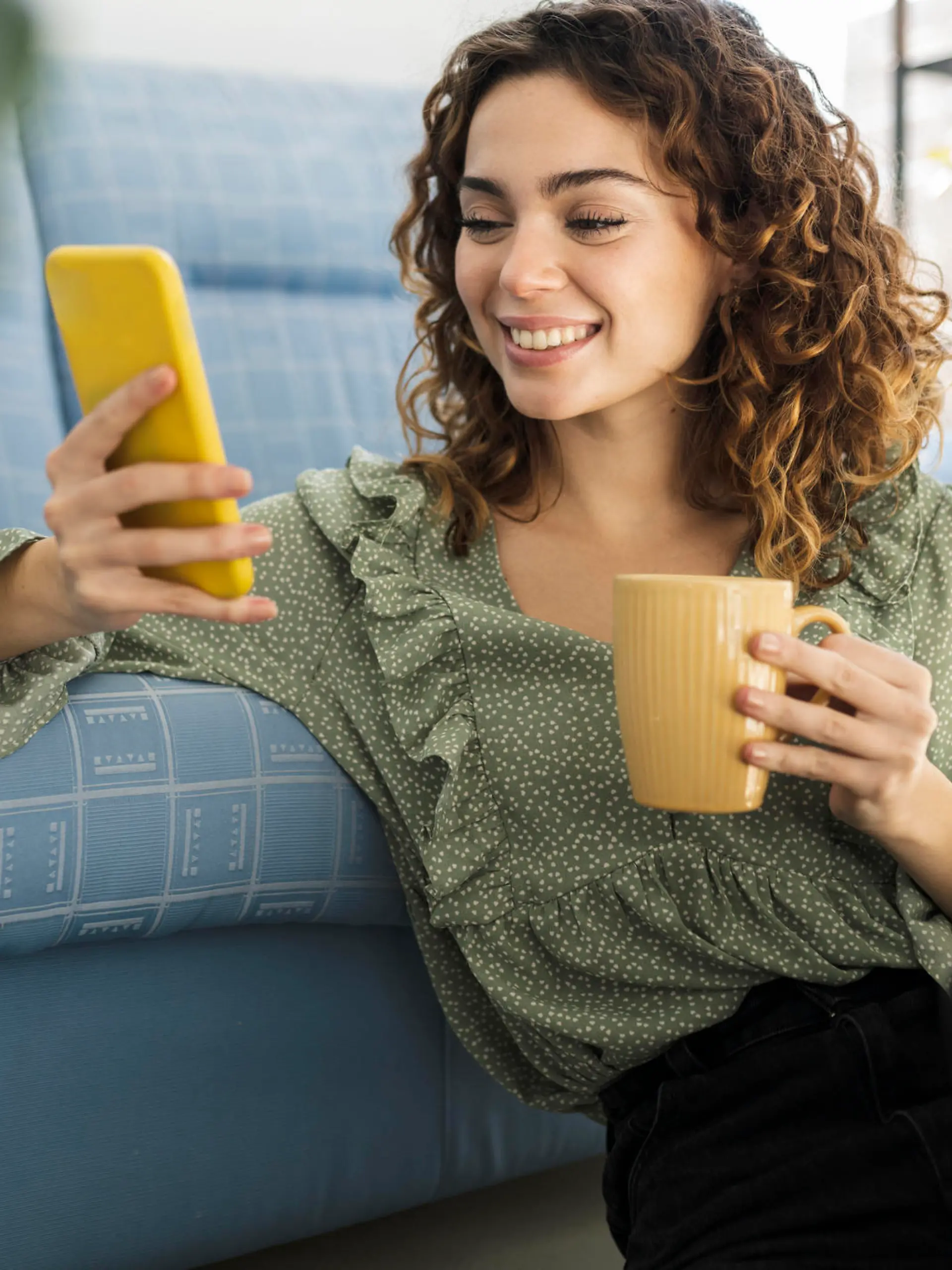 curly haired woman sitting on sofa with a cup of hot drink and mobile phone