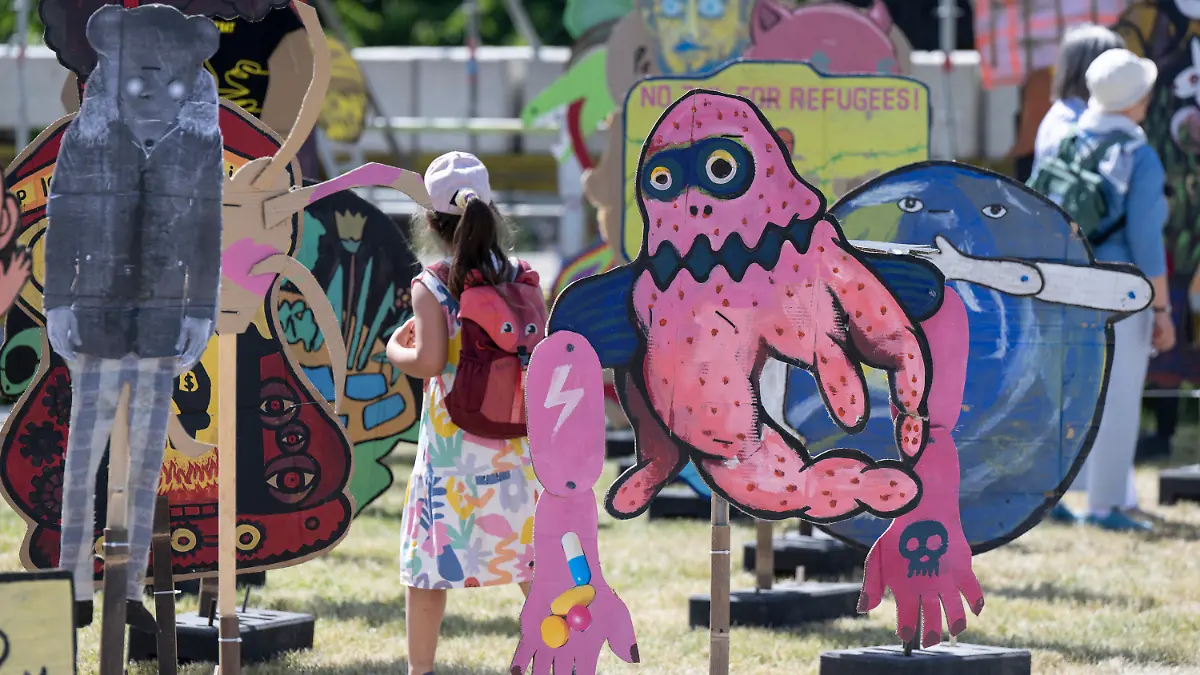17.06.2022, Hessen, Kassel: Besucher gehen zwischen riesigen Pappfiguren der indonesischen Künstlergruppe ·Taring Padi· auf dem Friedrichsplatz hindurch. Die internationale Kunstausstellung documenta fifteen ist zunächst für Fachbesucher geöffnet, ab dem 18. Juni 2022 steht sie allen Interessierten offen. Foto: Boris Roessler/dpa - ACHTUNG: Nur zur redaktionellen Verwendung im Zusammenhang mit einer Berichterstattung über die documenta 15 und nur mit vollständiger Nennung des vorstehenden Credits +++ dpa-Bildfunk +++