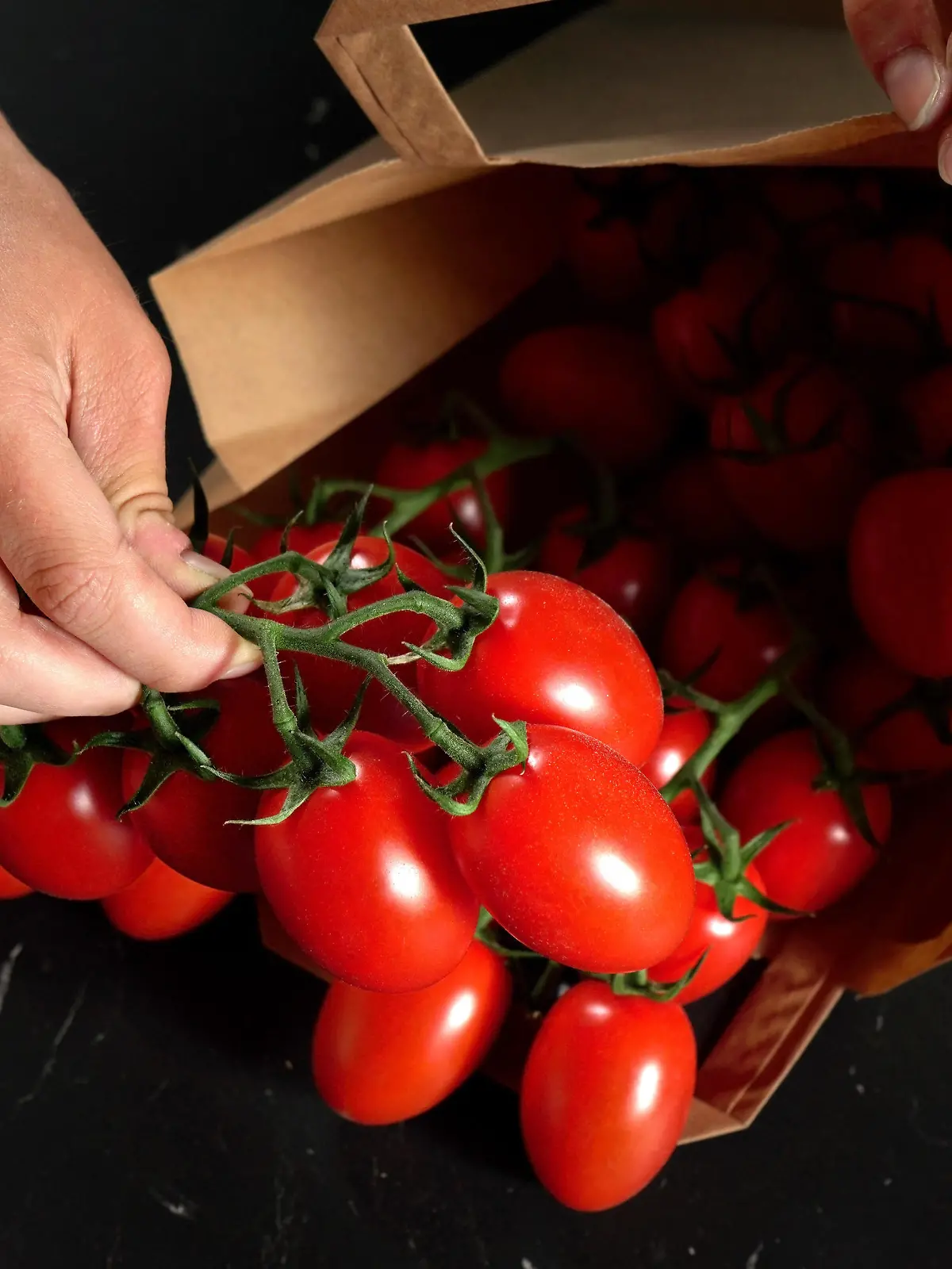 Detail on hands getting small red cherry tomatoes with green vines from brown paper bag, over dark black marble board.