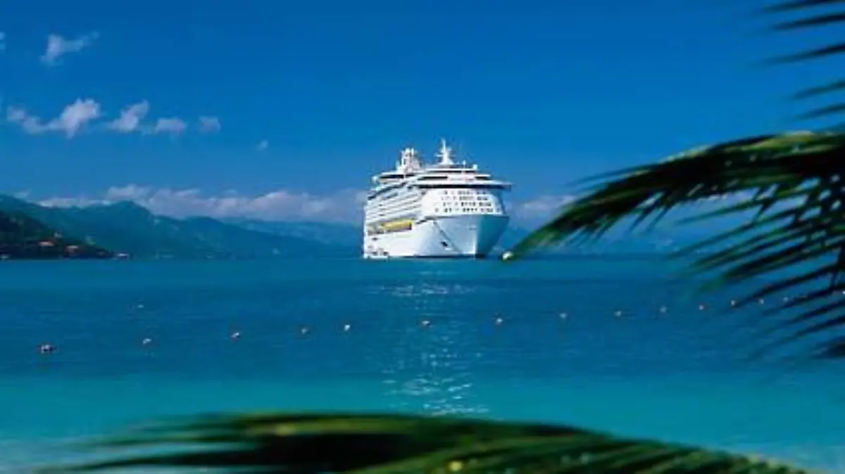 Kreuzfahrtschiff ankert in einer Bucht in der  Karibik, Palmenblaetter im Vordergrund.