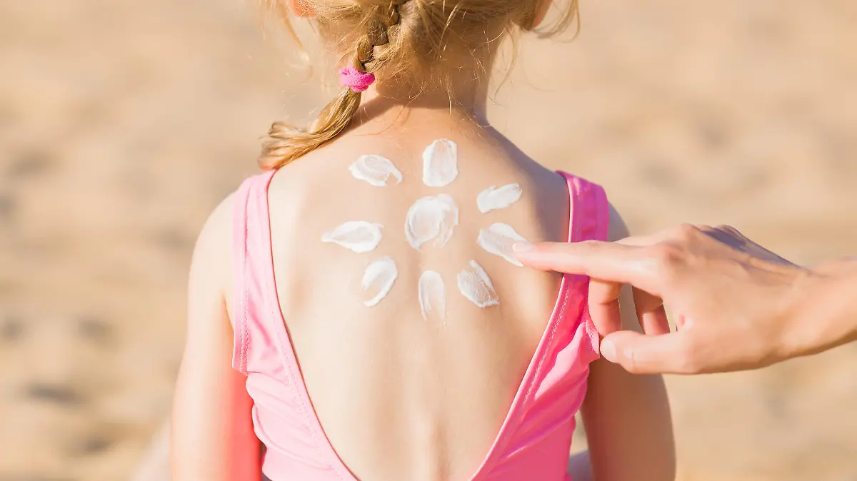 Young adult mother hand applying sunscreen lotion on little girl back. Sun shape created from white cream. Skin protection. Safety sunbathing in hot sunny day at beach. Rear view. Closeup.