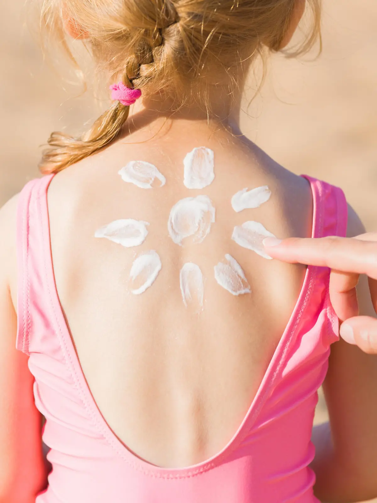 Young adult mother hand applying sunscreen lotion on little girl back. Sun shape created from white cream. Skin protection. Safety sunbathing in hot sunny day at beach. Rear view. Closeup.